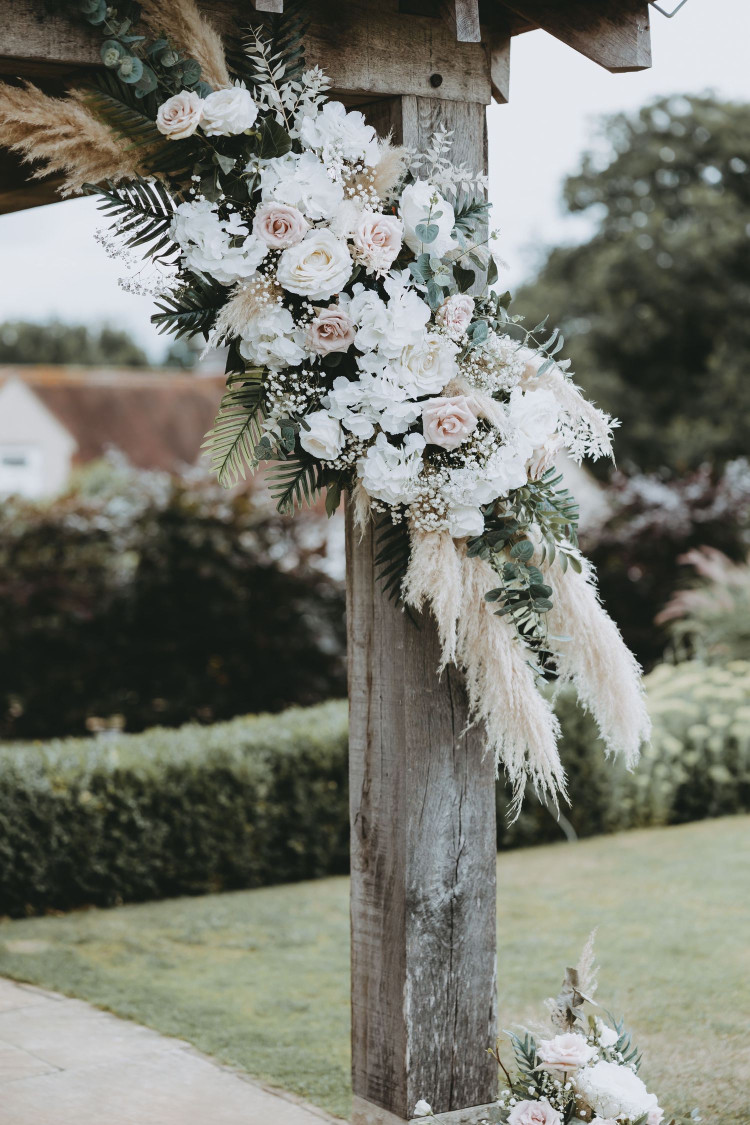 Rustic wooden posts adorned with white flowers and dried pampas grass create an elegant outdoor wedding ceremony space.