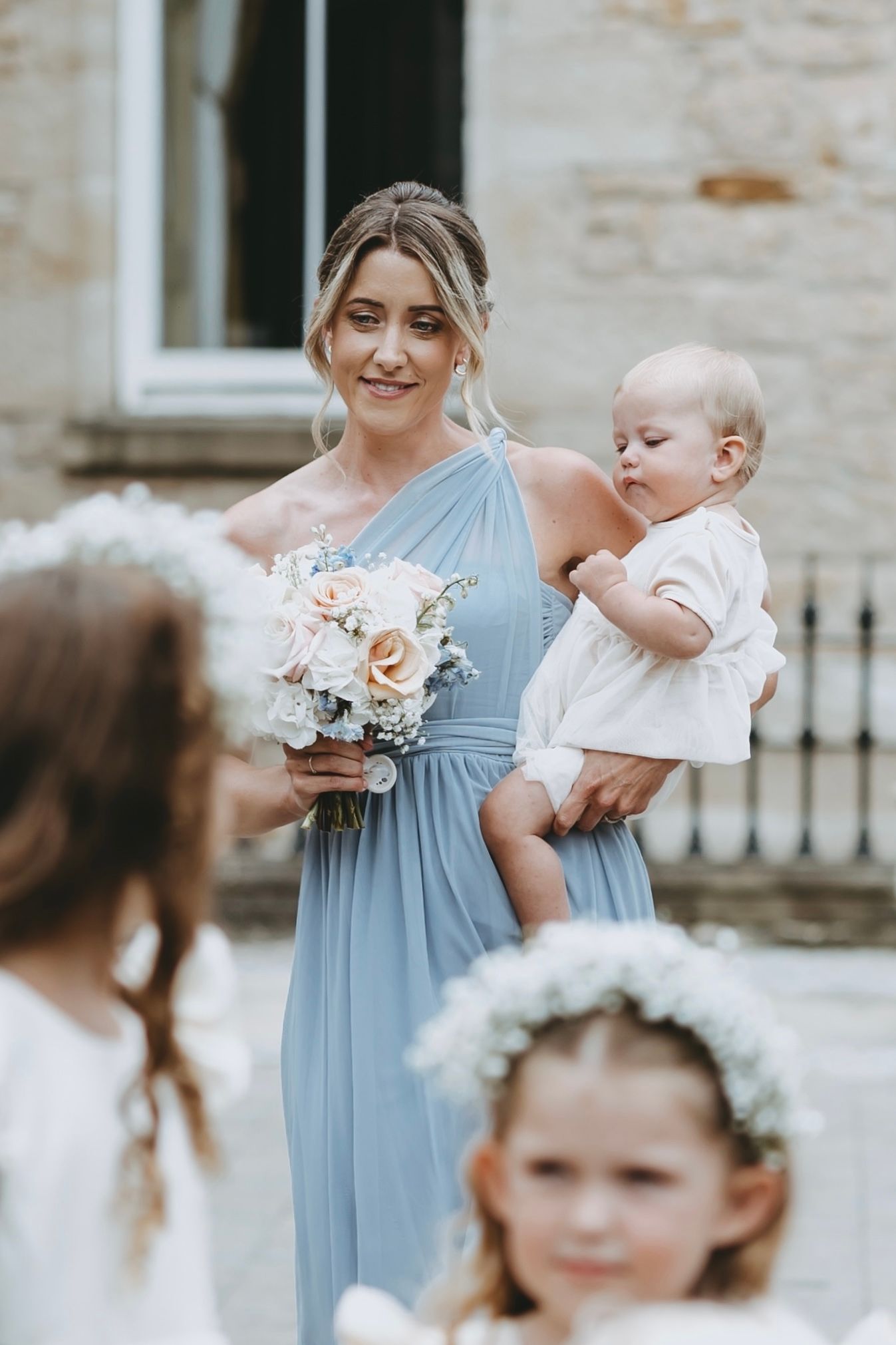 Bridesmaid in flowing powder blue dress holds a baby and bouquet at outdoor wedding ceremony.