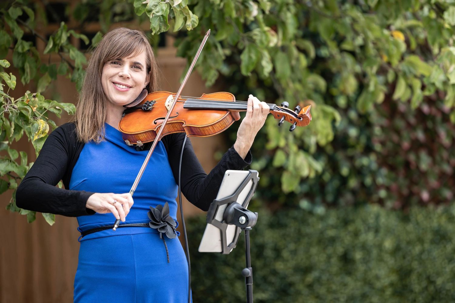 Violinist in bright blue dress performs outdoors with greenery in background.