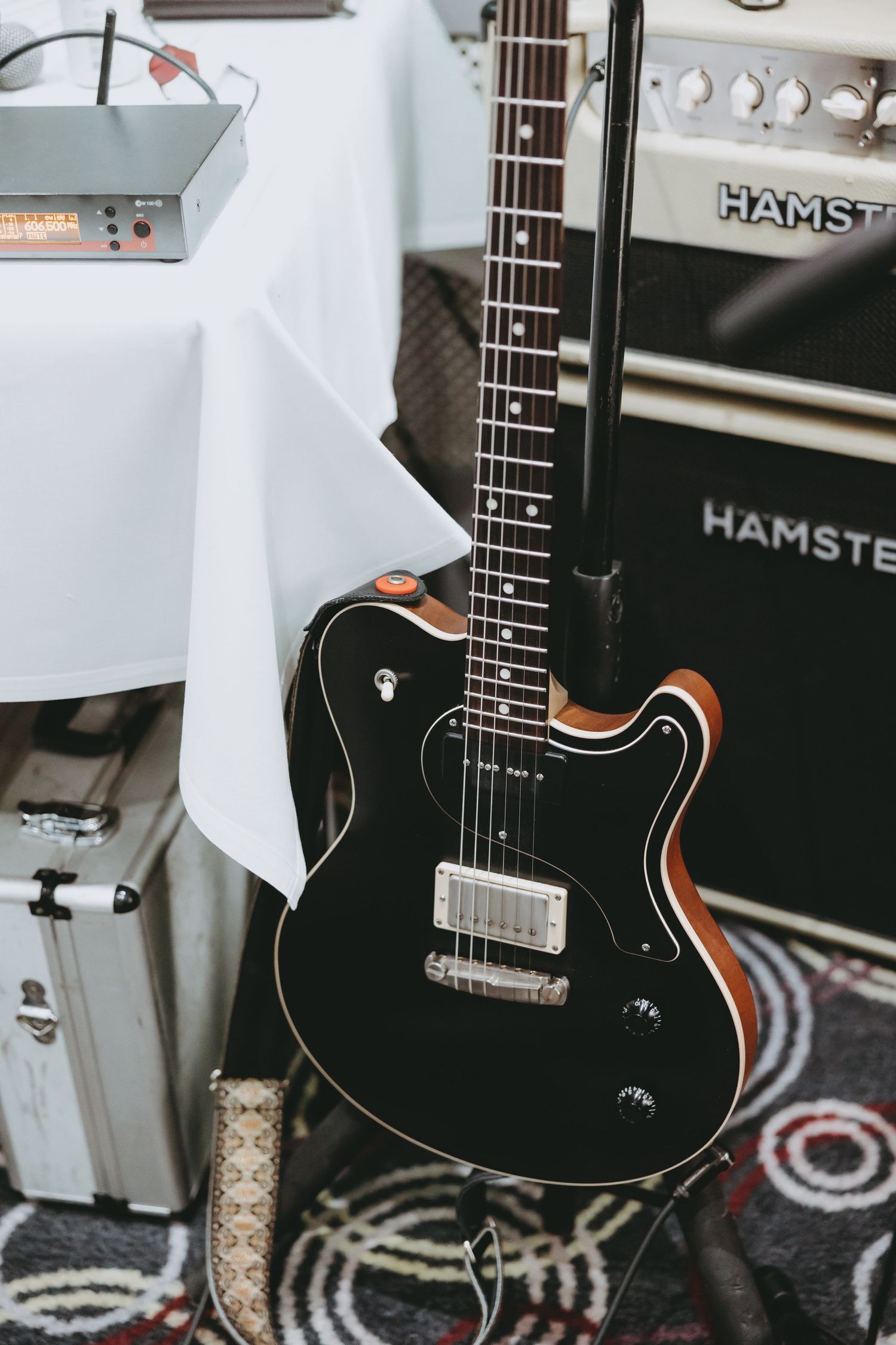 Black electric guitar with orange accents sits against a Hamster amplifier on a patterned carpet.