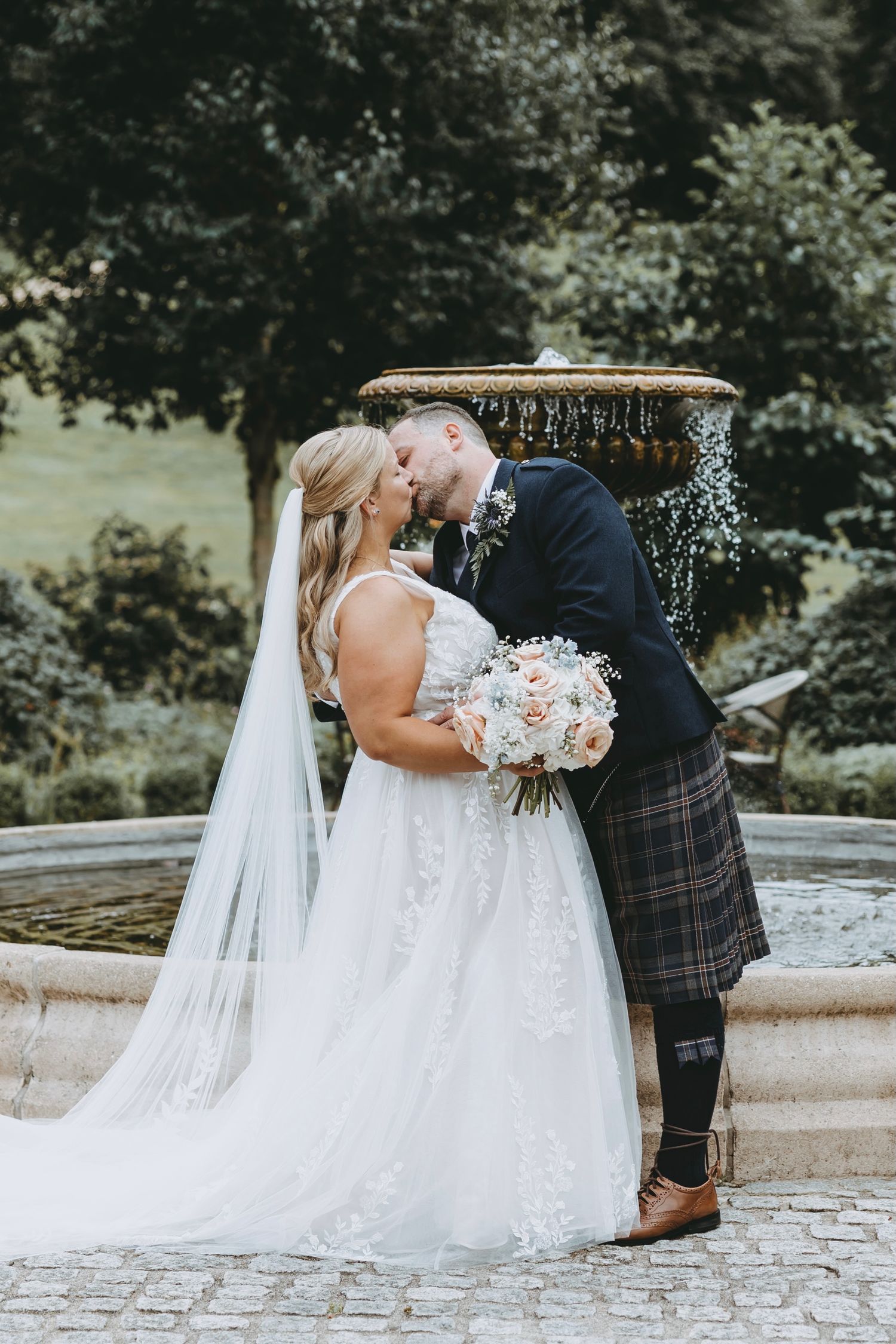 Couple shares intimate moment on stone steps with bride in flowing white gown and groom in Scottish attire.