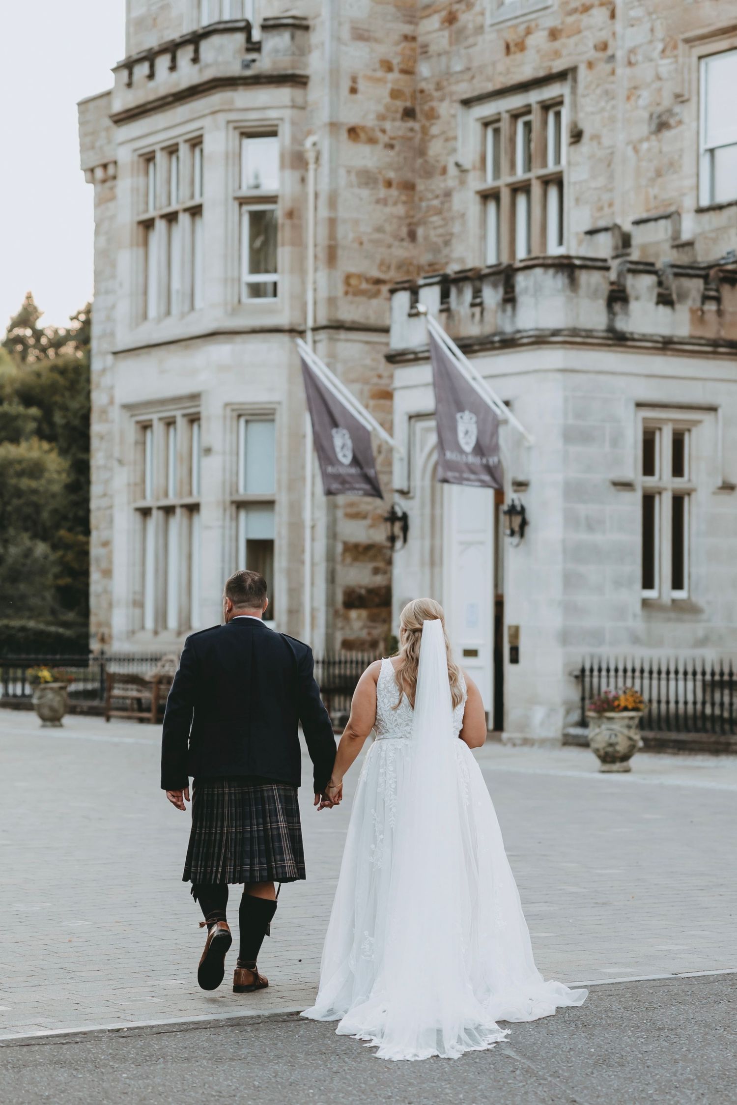 Newlyweds walking away from stone castle venue with bride in flowing white gown.