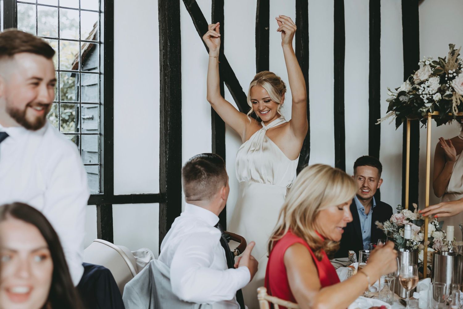Wedding reception guests cheer and raise glasses during toasts in a timber-framed venue.