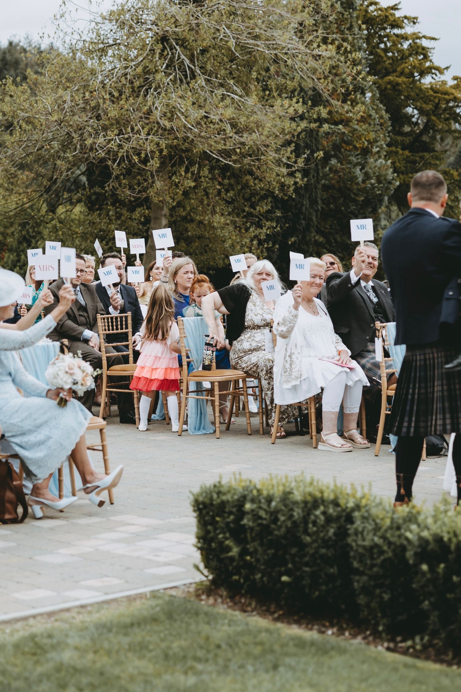 Wedding guests seated in wooden chairs participate in an interactive game with white signs.