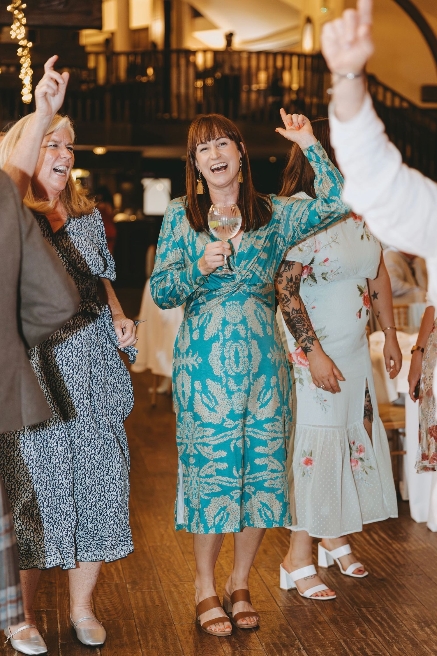Guests in patterned teal and floral dresses dancing together at a wedding reception.