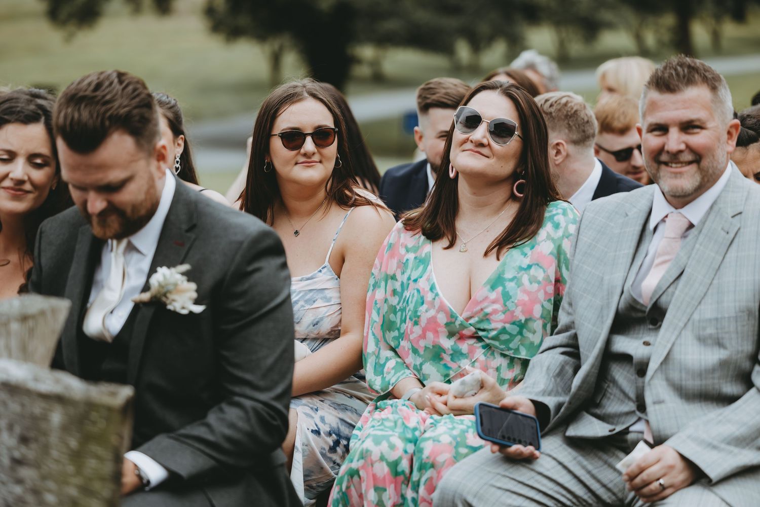 Wedding guests in summer attire seated on wooden benches during an outdoor ceremony.