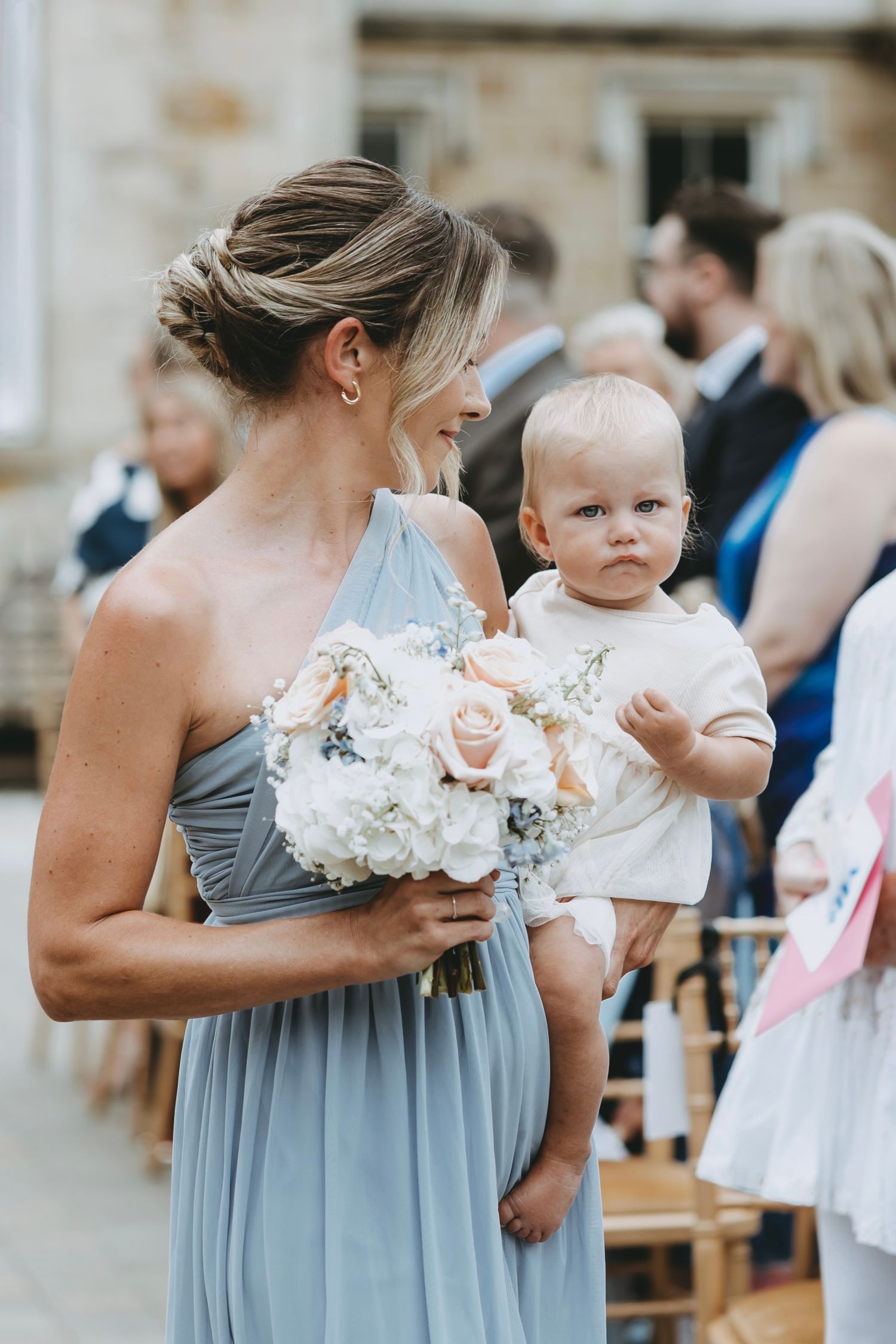 Wedding attendees in pale blue dresses holding floral bouquets gather outside.