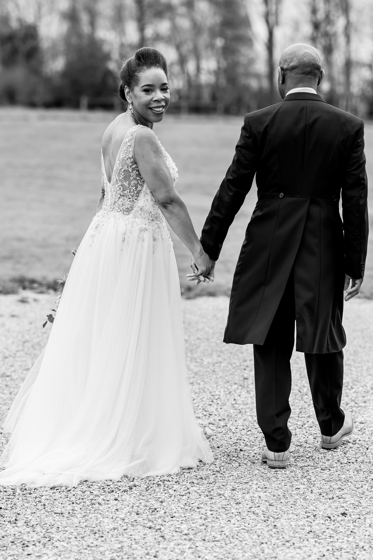 Newlyweds walking hand in hand on gravel path in elegant wedding attire.