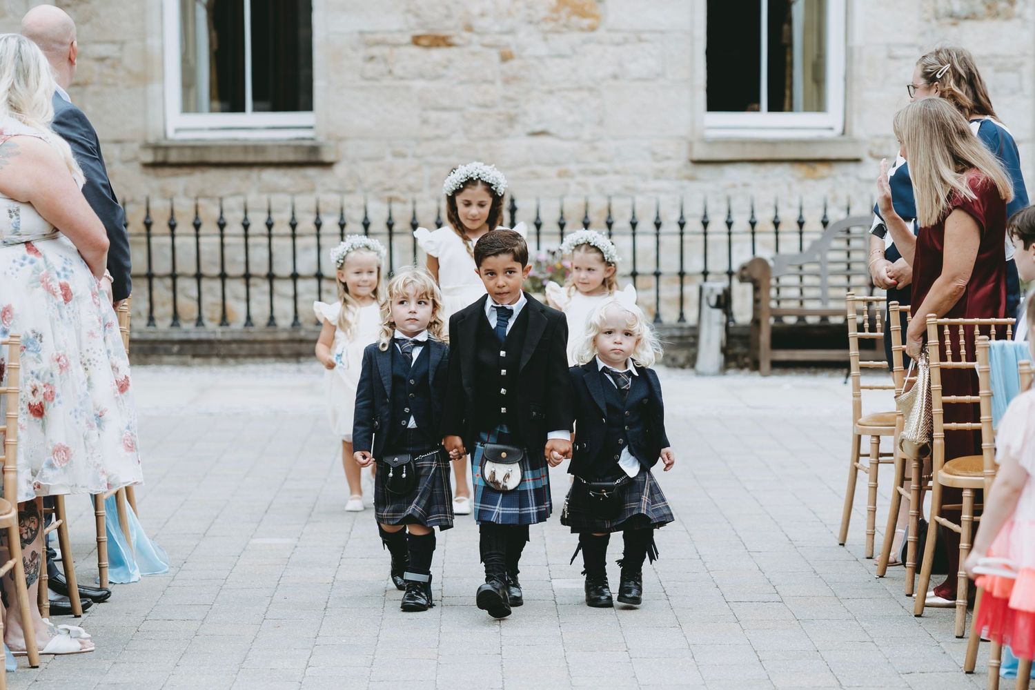 Children in traditional Scottish wedding attire walking down aisle at stone building venue.