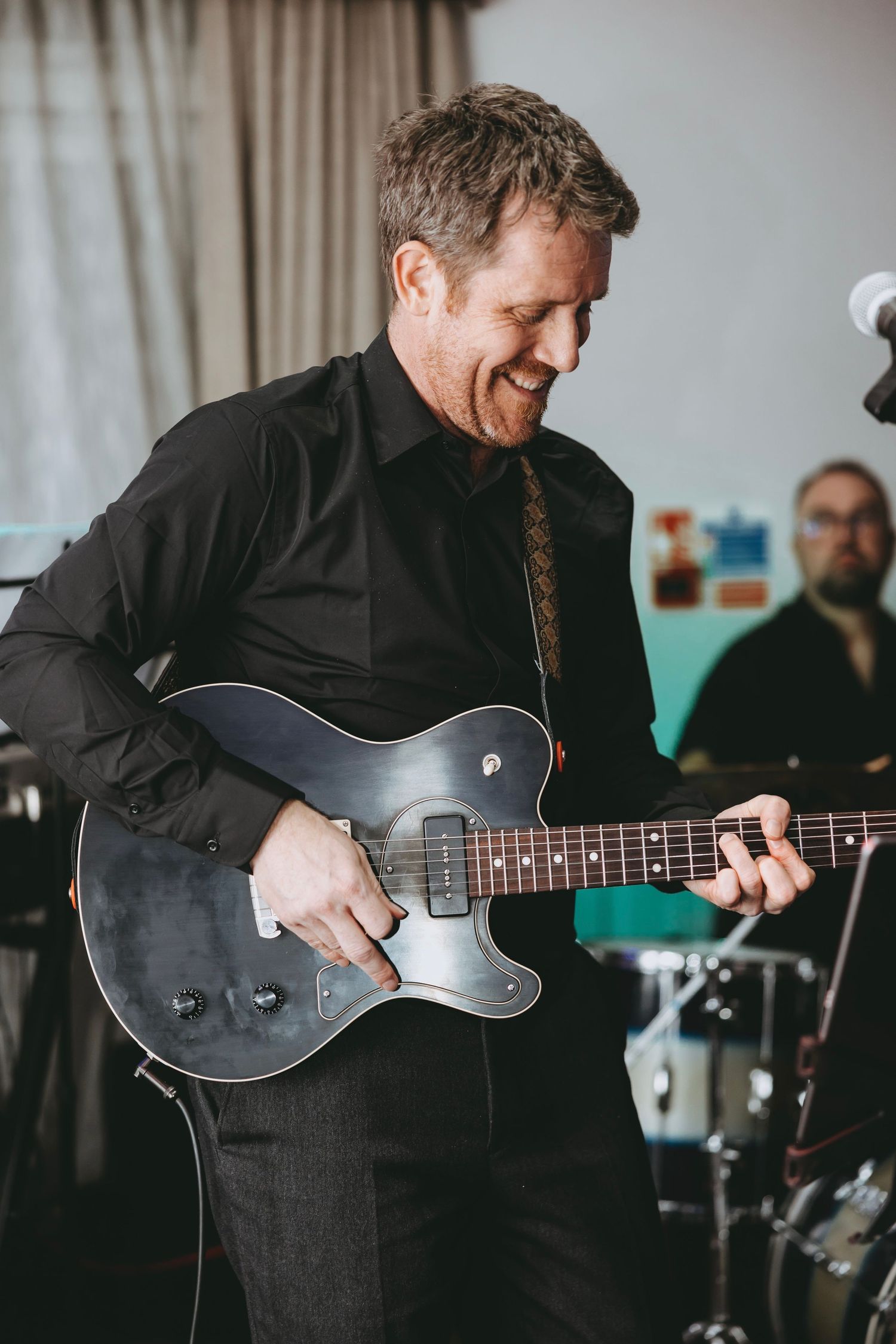 Musician in black suit plays dark electric guitar during indoor performance session.