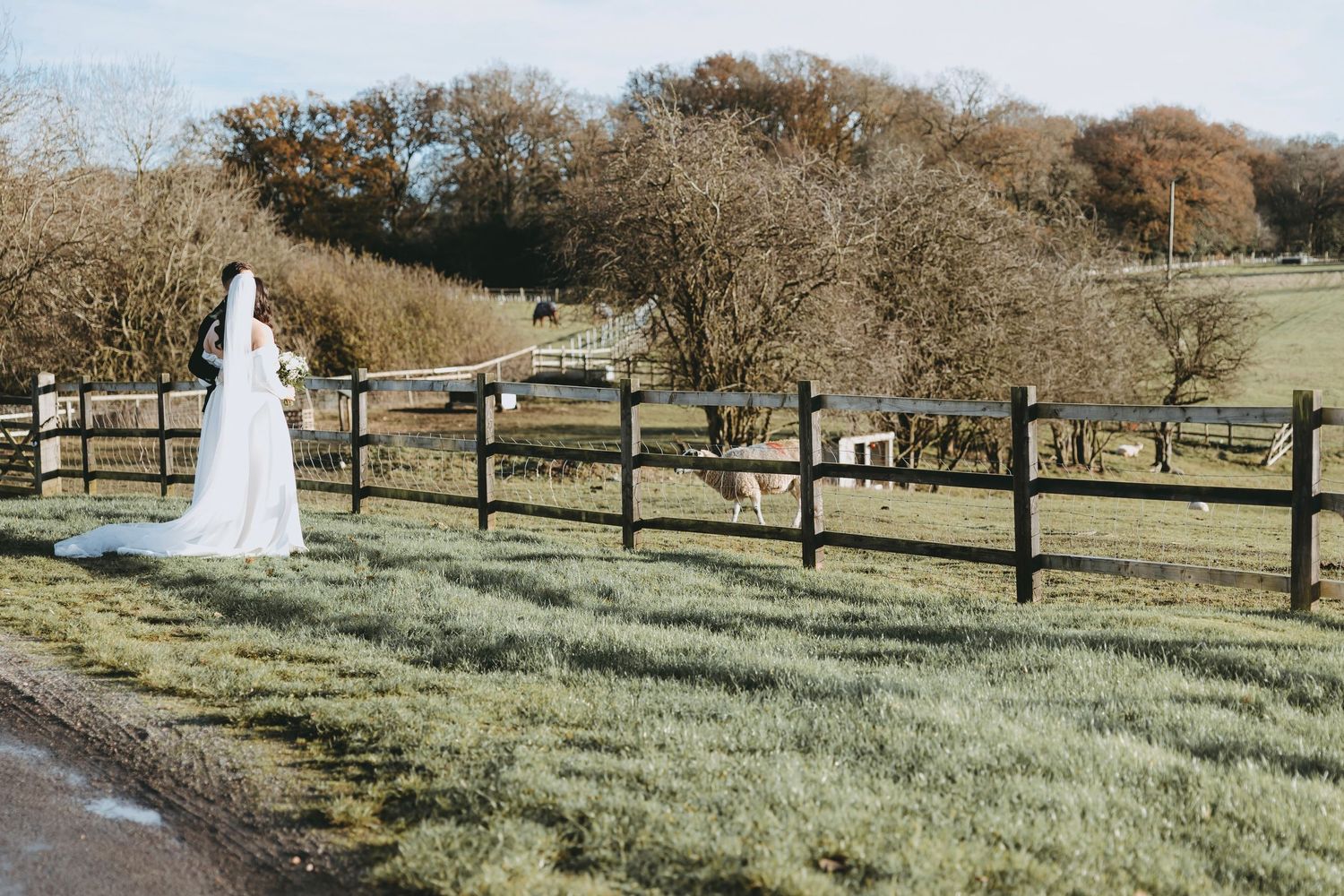 A person in a white wedding dress walks along a rustic wooden fence in a scenic outdoor field setting.