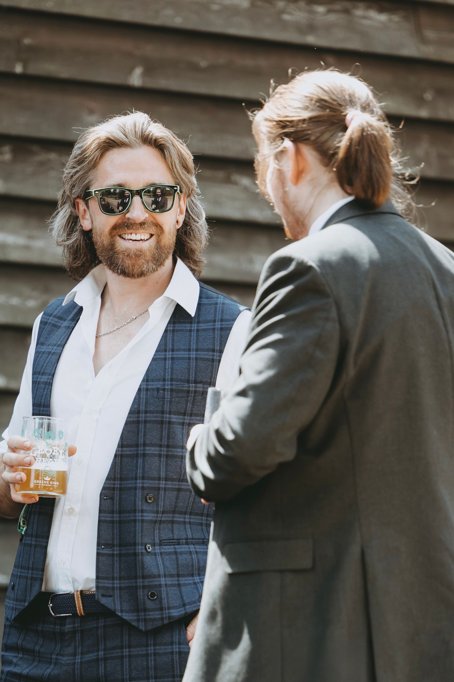 Wedding guest in plaid vest and sunglasses smiles while holding a drink at outdoor reception.