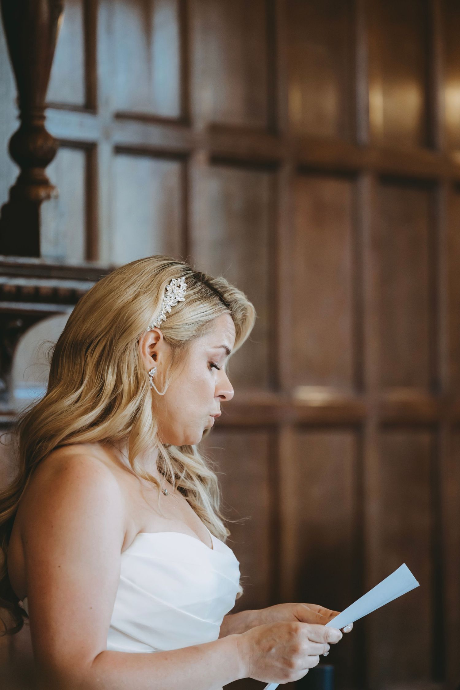 A person in white reads from a paper in front of ornate wooden paneling.