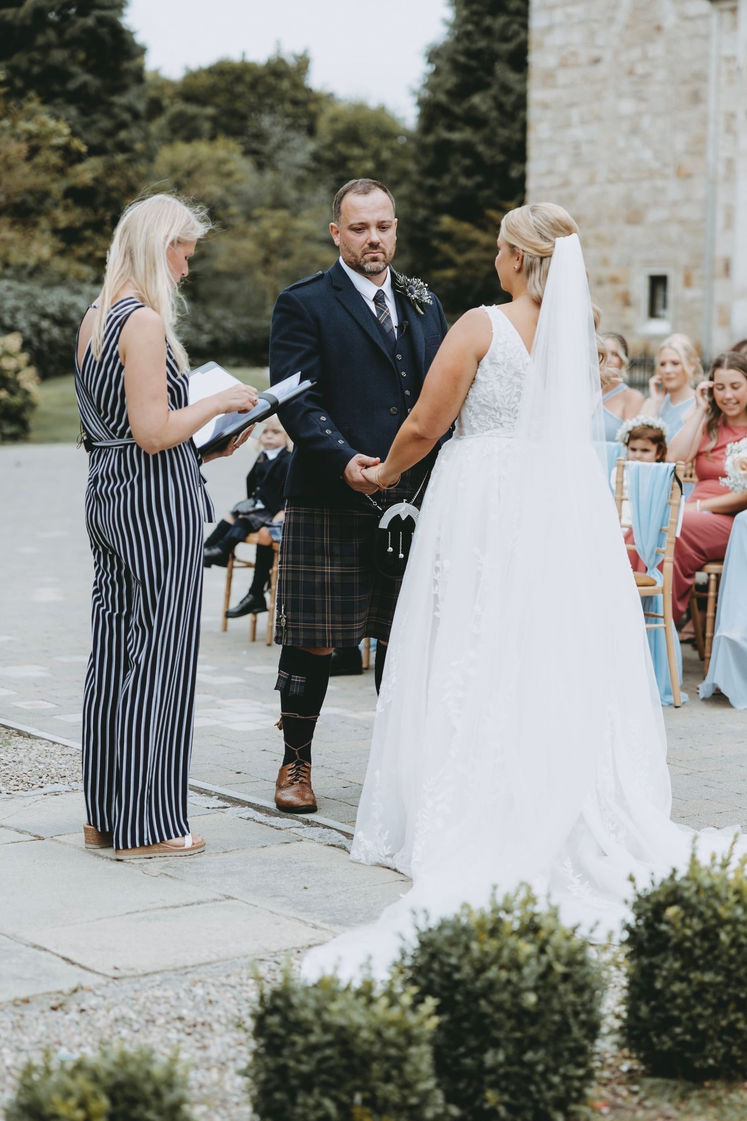Wedding ceremony in progress outside a stone building with guests seated on white chairs.