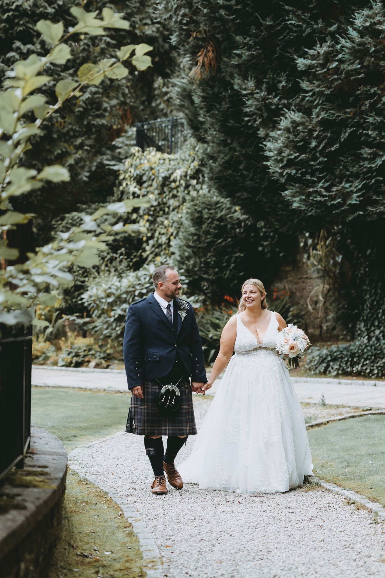 Couple in wedding attire sharing moments on a garden path with evergreen backdrop.