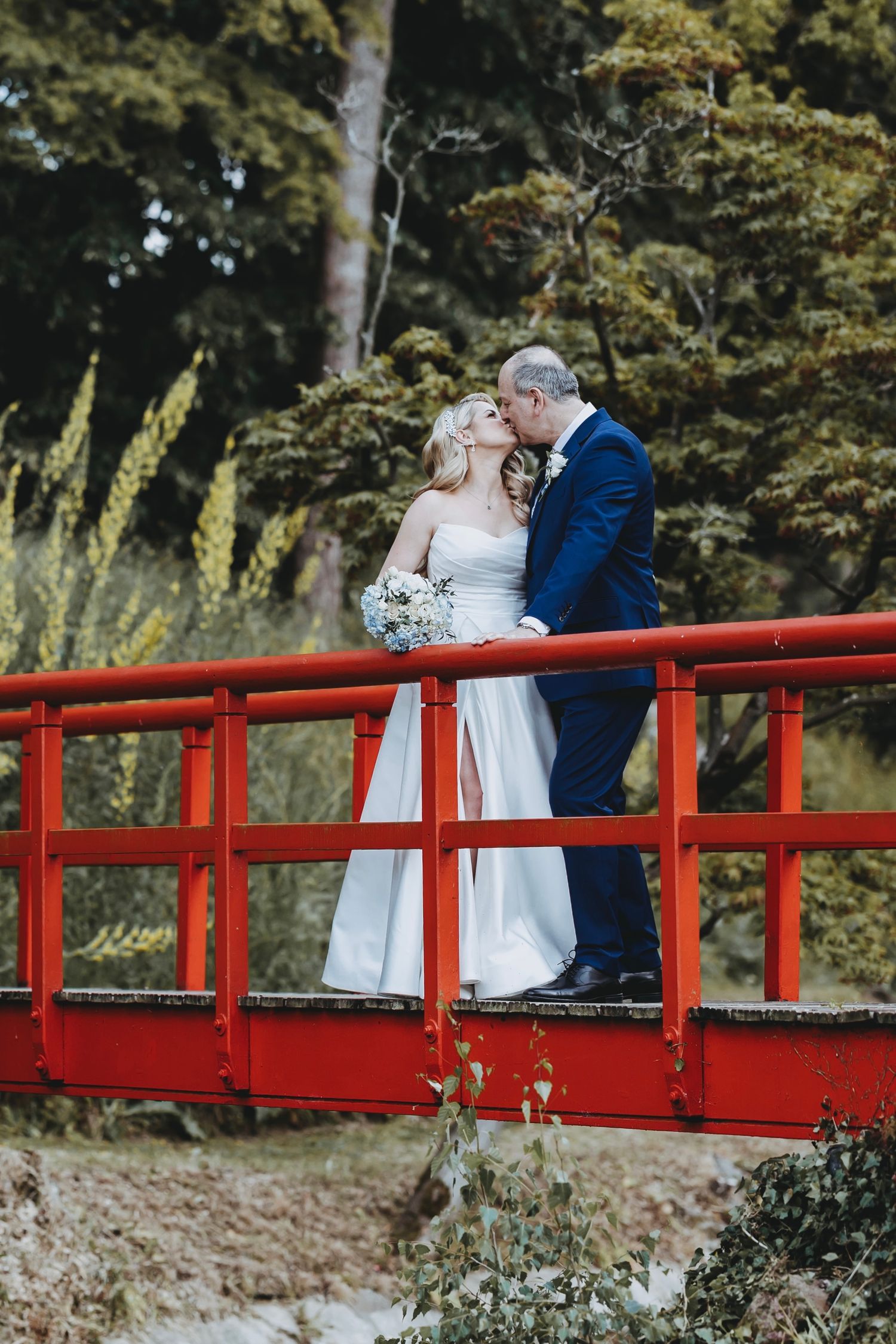 A couple in wedding attire stands on a bright red bridge surrounded by natural landscaping.