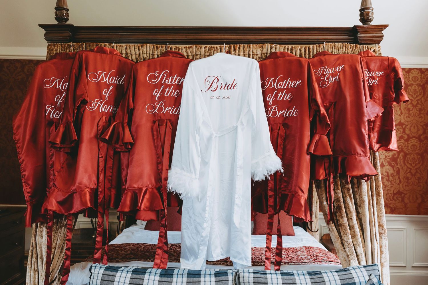 White bridal robe with feather trim displayed among red bridesmaid robes on a bed, perfect for wedding party getting ready photos.