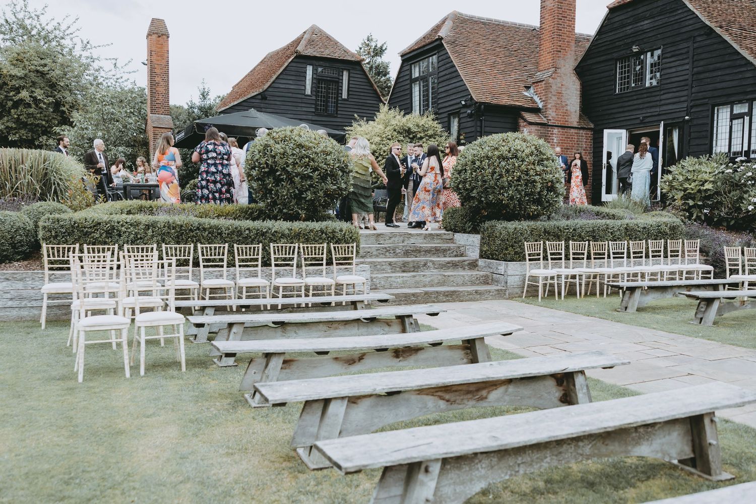 White wooden chairs arranged in rows with rustic wooden benches at an outdoor wedding ceremony space.
