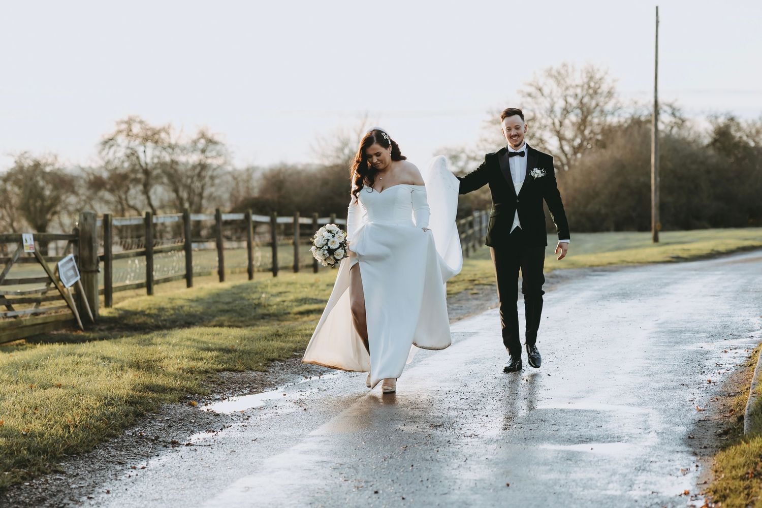 A couple walks along a wet path surrounded by green fields on their wedding day.
