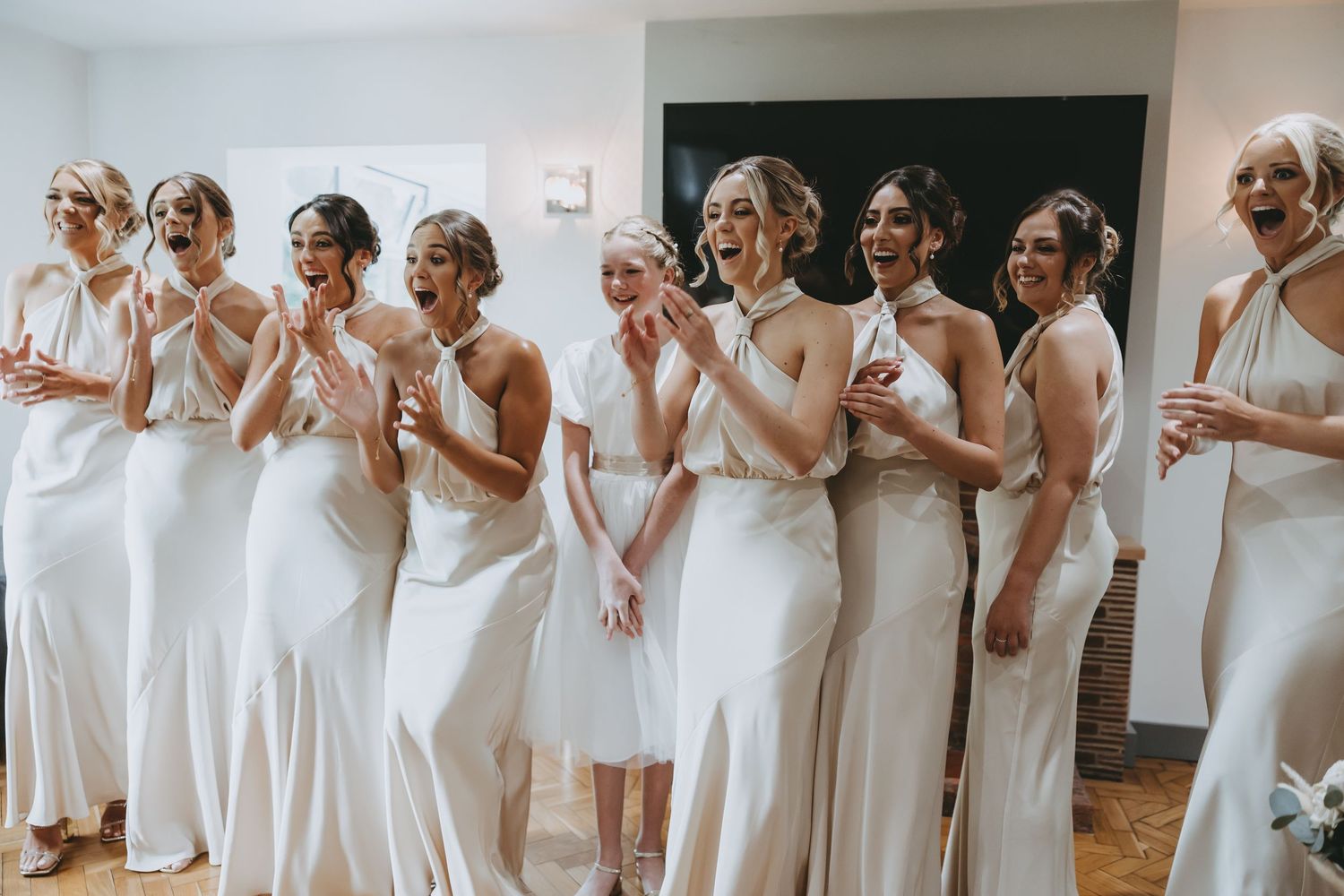 Row of bridesmaids in flowing white dresses reacting with excitement during wedding preparations.