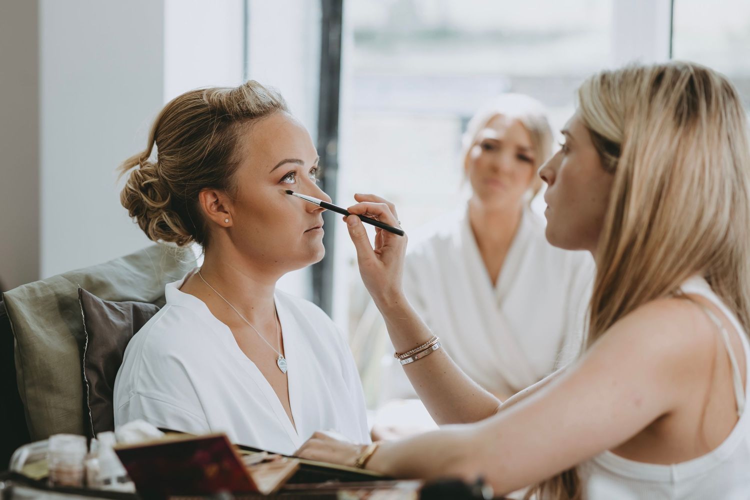 Bridal party getting ready with makeup application in bright natural light setting.