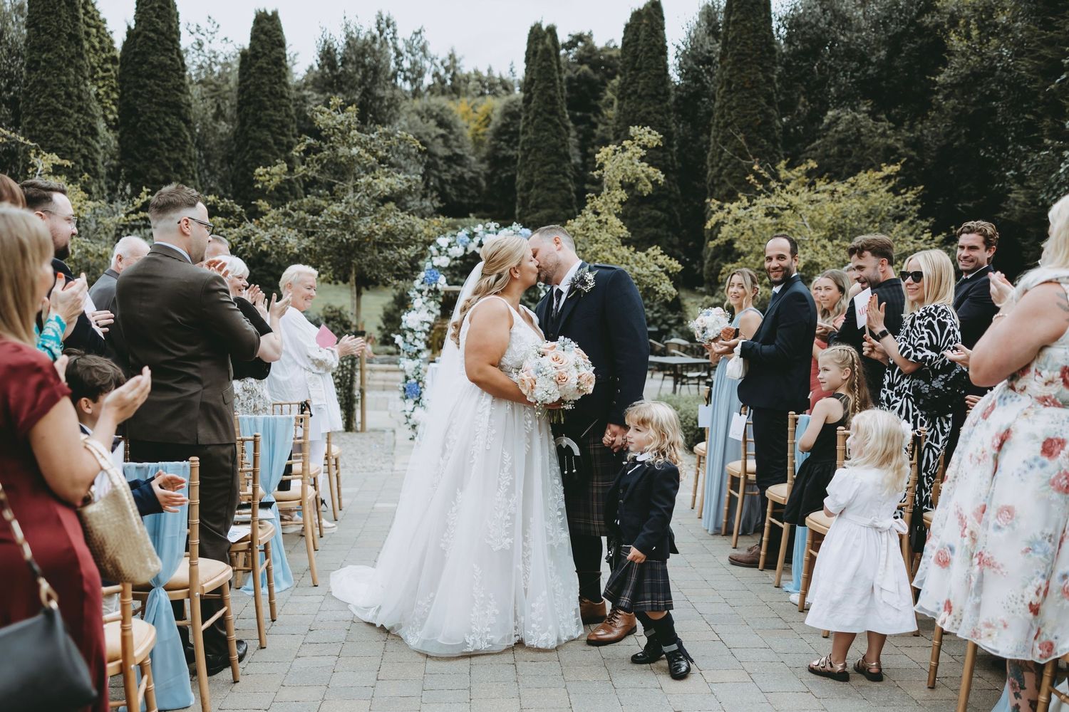 Wedding guests seated on wooden chairs during an outdoor ceremony with trees in background.