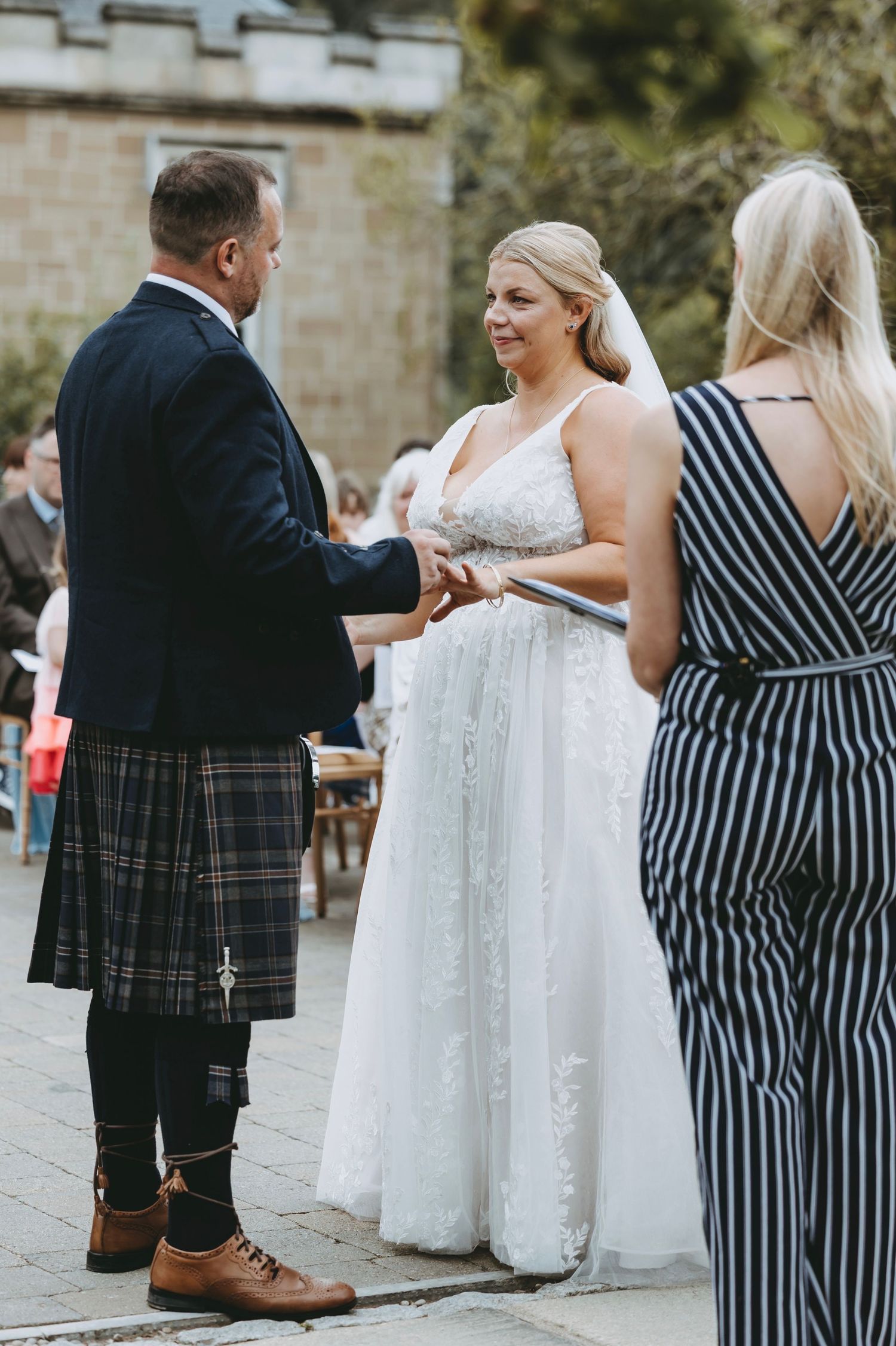Outdoor wedding ceremony with guests in striped dress and traditional Scottish attire.