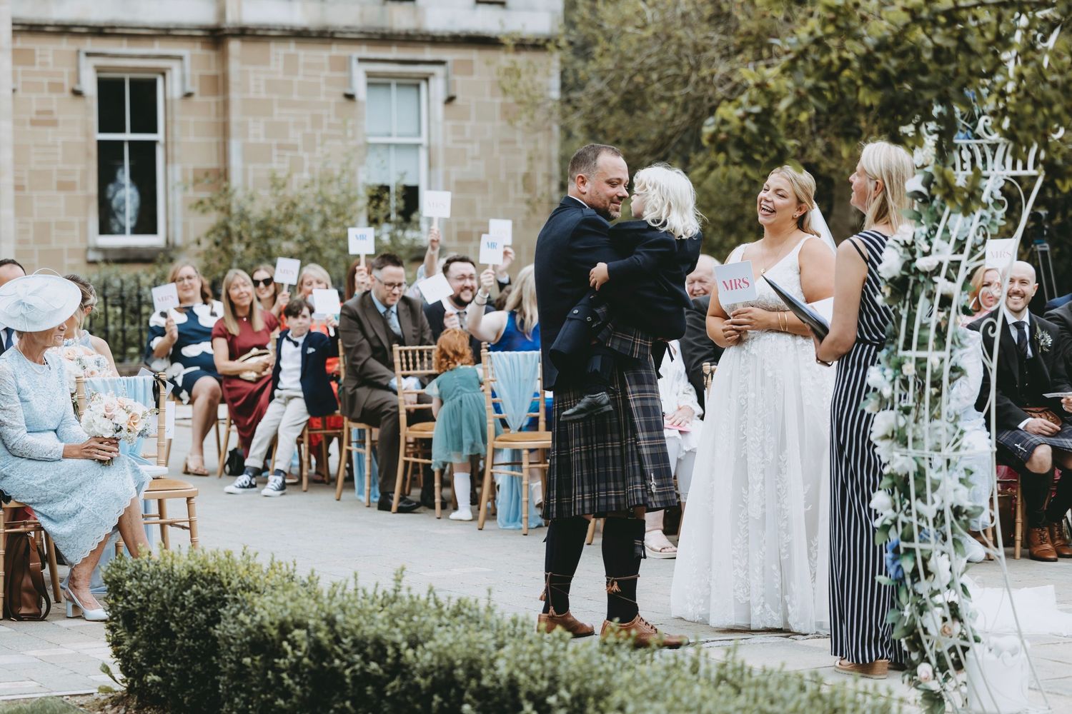 Outdoor wedding at stone mansion with decorative metal arch and seated guests.
