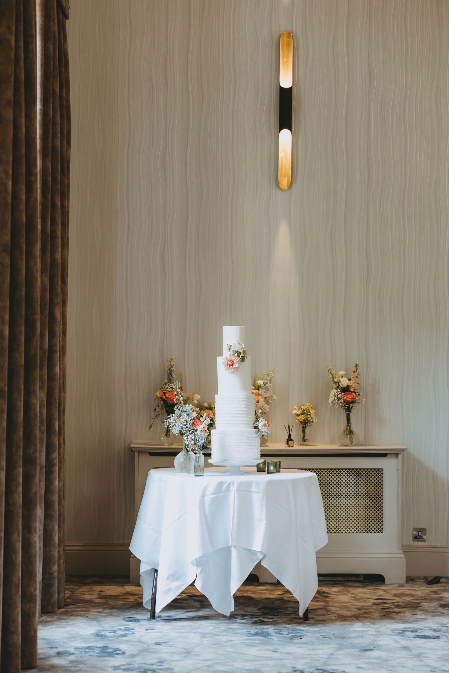 Elegant three-tier white wedding cake with floral accents on a draped table, flanked by flower arrangements in a softly lit room.