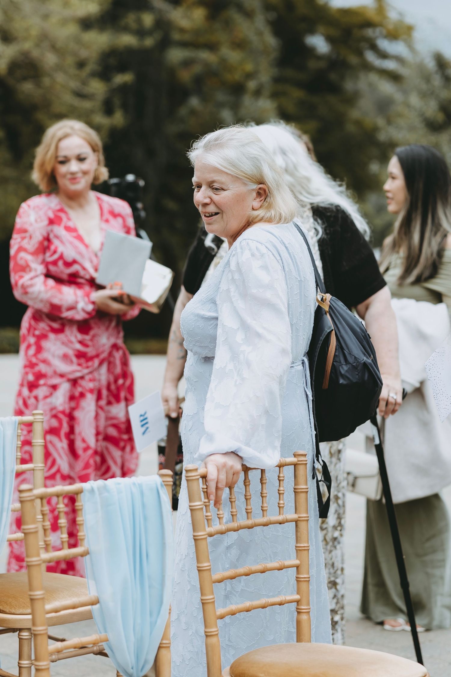 Wedding guests mingling around wooden chairs decorated with light blue sashes outdoors.