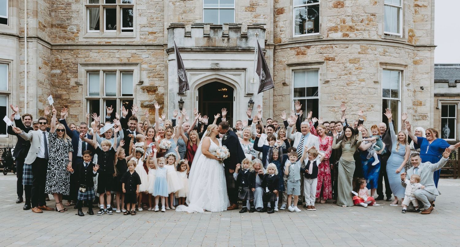 Large group wedding photo of guests gathered in front of historic stone building, Crossbasket Castle, Scotland.