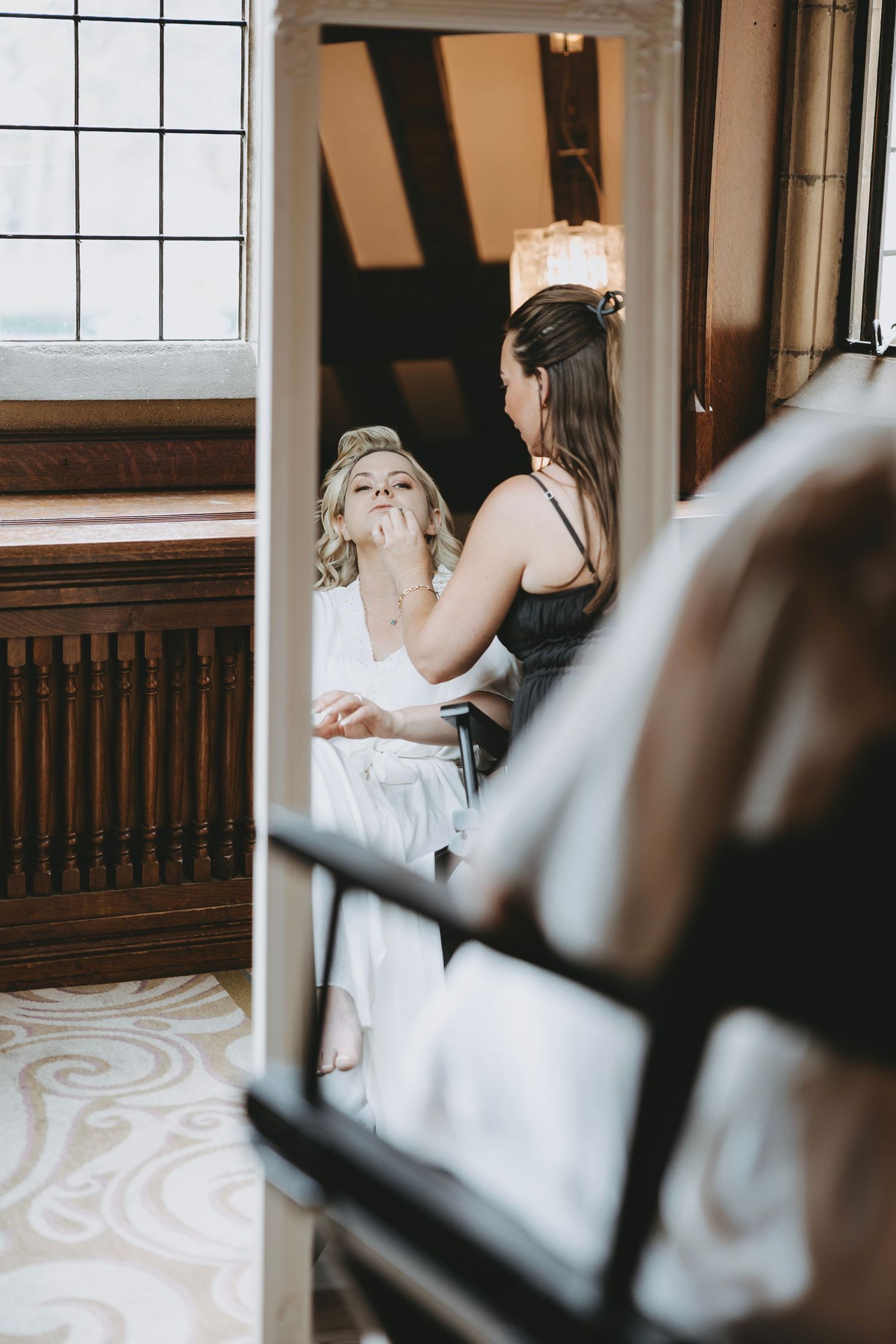 Two people reflected in a mirror during a makeup session in an elegant room.