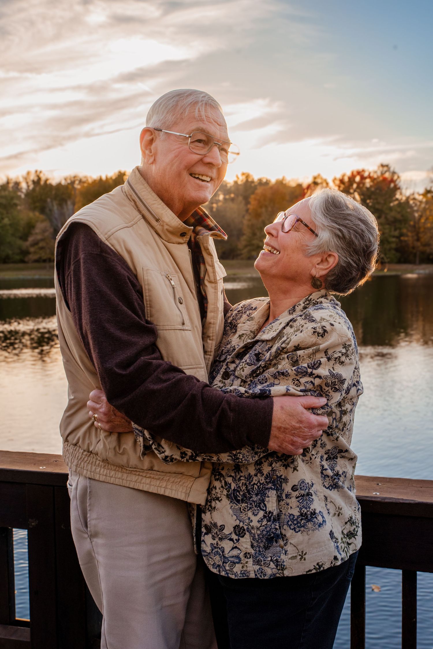 Elderly couple laughing and embracing on a dock by a lake at sunset.