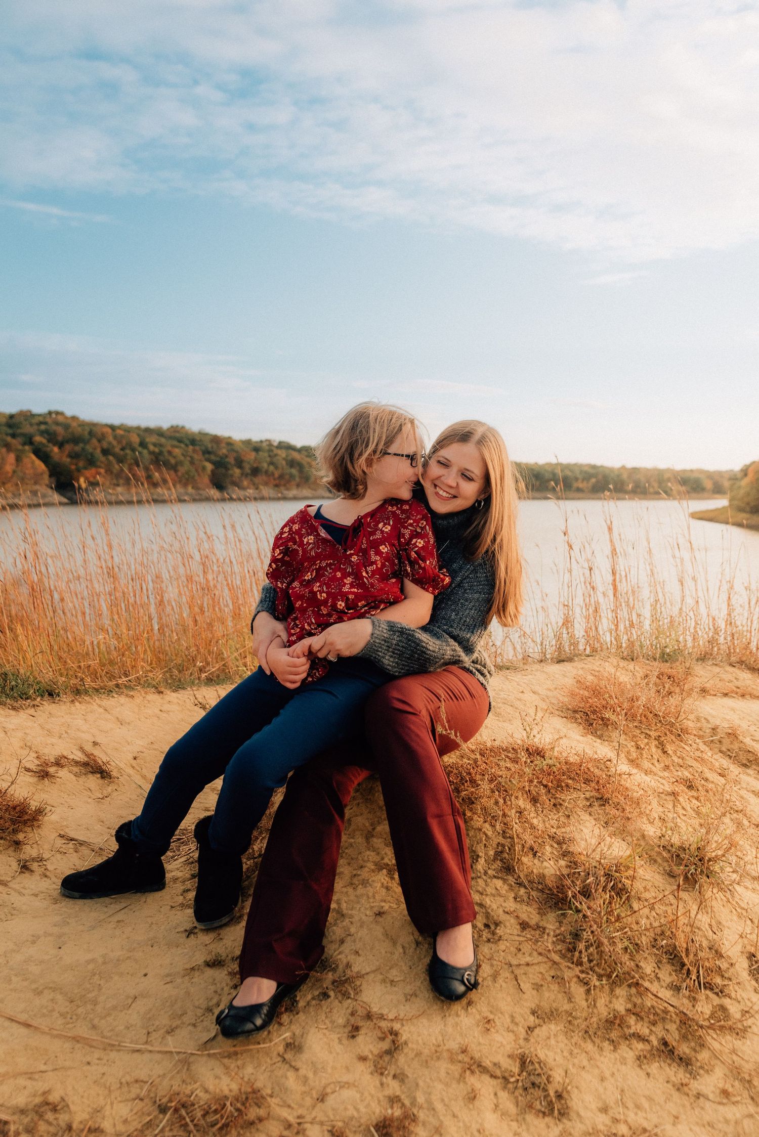 Two people sitting together on sandy beach cliff at sunset with tall grass in background.