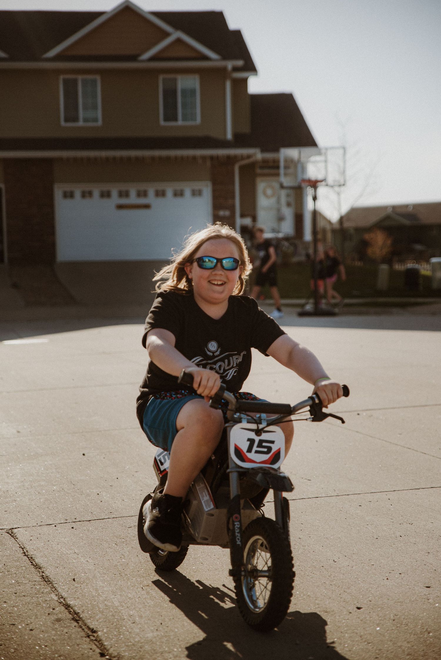 Child riding a small motorcycle in a suburban driveway wearing sunglasses and casual attire.