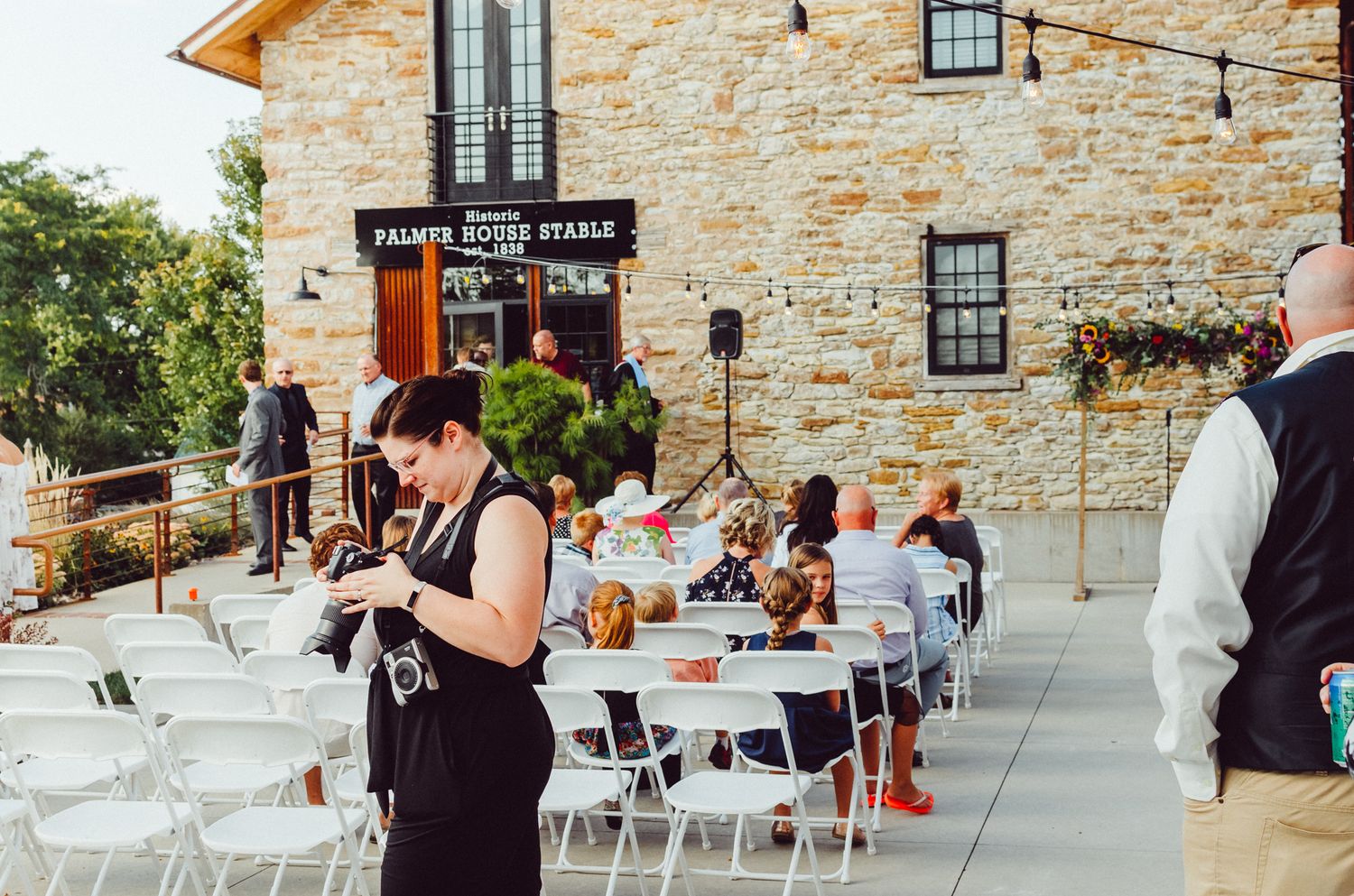 Photographer looking at her camera in front of a stone building exterior with outdoor seating area and string lights.