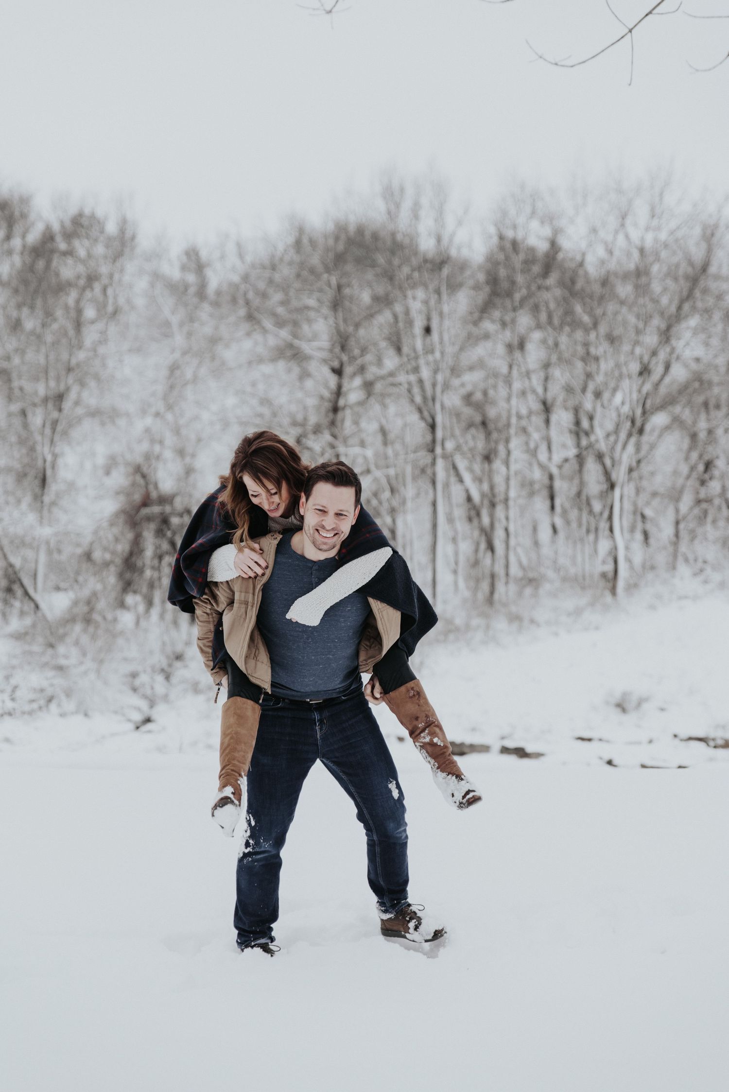 Two people playing and laughing together in a snowy winter landscape with bare trees in the background.