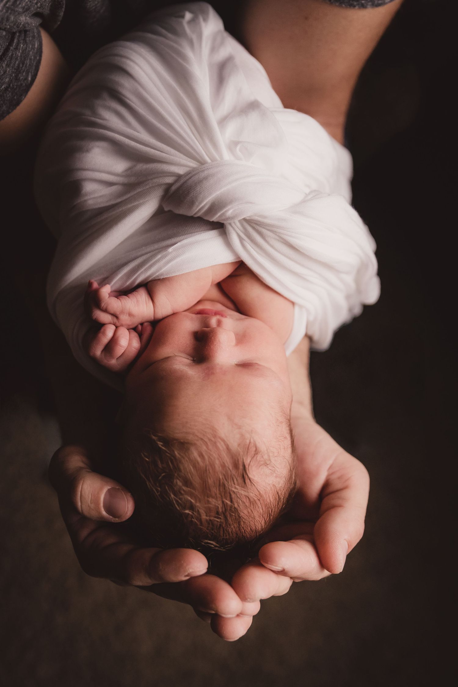 Artistic close-up of a sleeping newborn wrapped in white fabric against dark background.