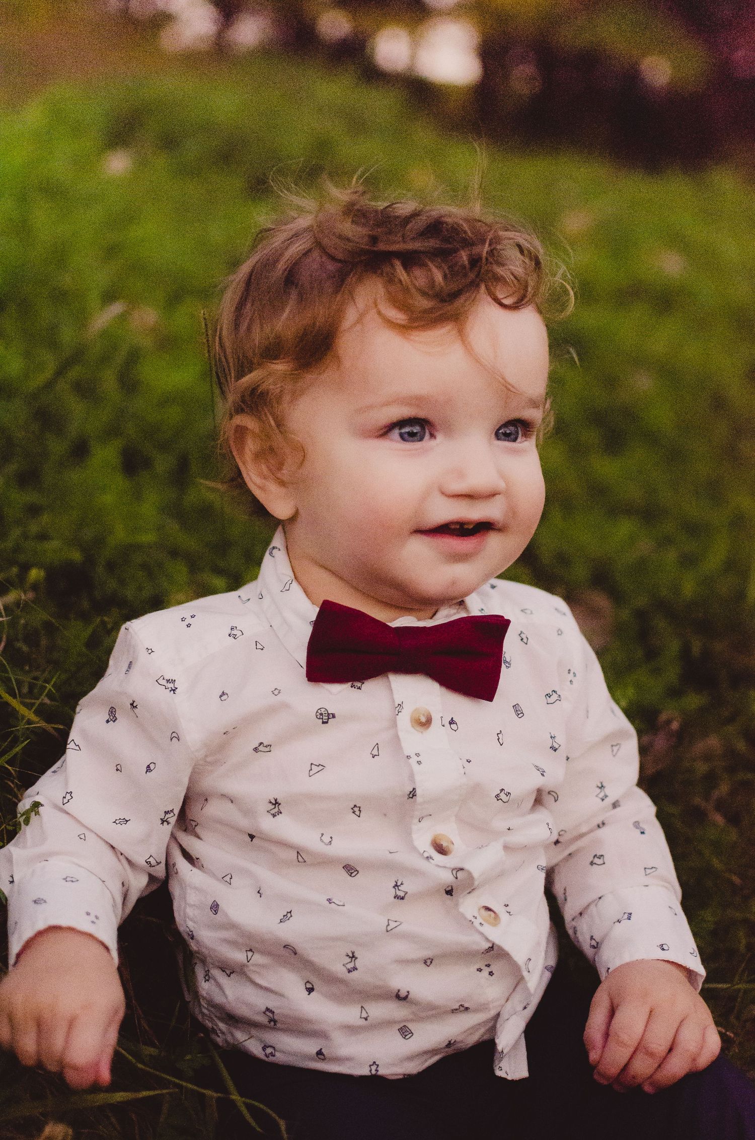 Baby wearing white patterned shirt and burgundy bow tie sitting on grass.