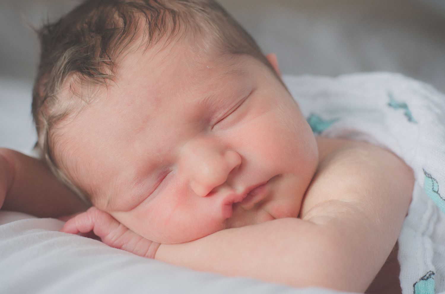Sleeping newborn baby photographed in soft natural light against white bedding.