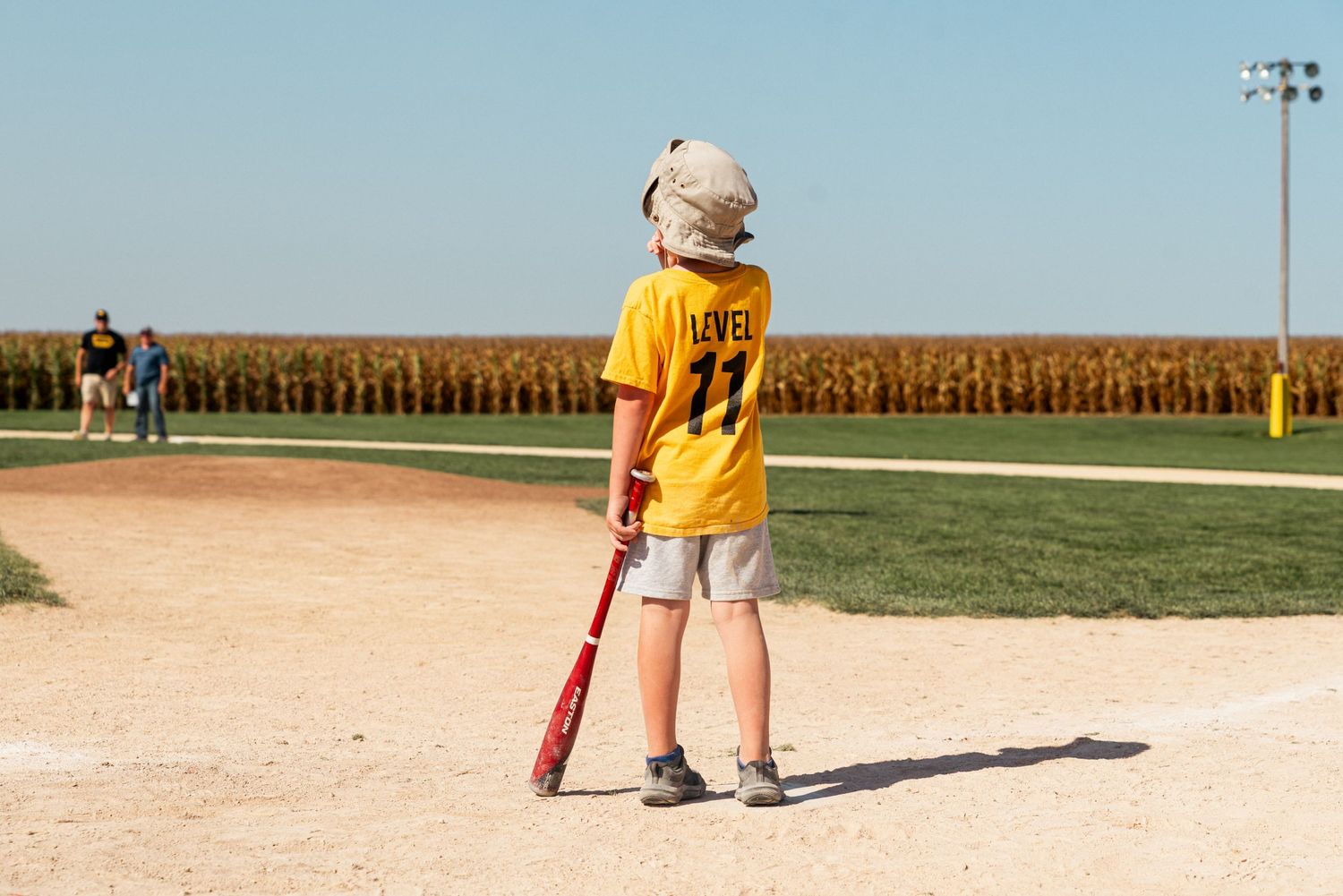 Baseball players on field with cornfield backdrop during sunny day.