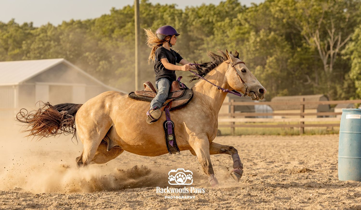 Young girl in purple helmet rides a palomino horse at full gallop on a dirt arena during a barrel racing event.