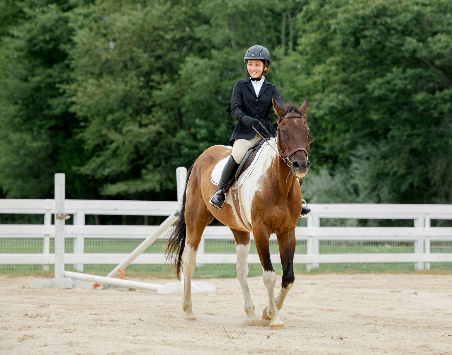 Young equestrian rider in black jacket and helmet riding a chestnut pinto horse in a sandy arena with white fence.