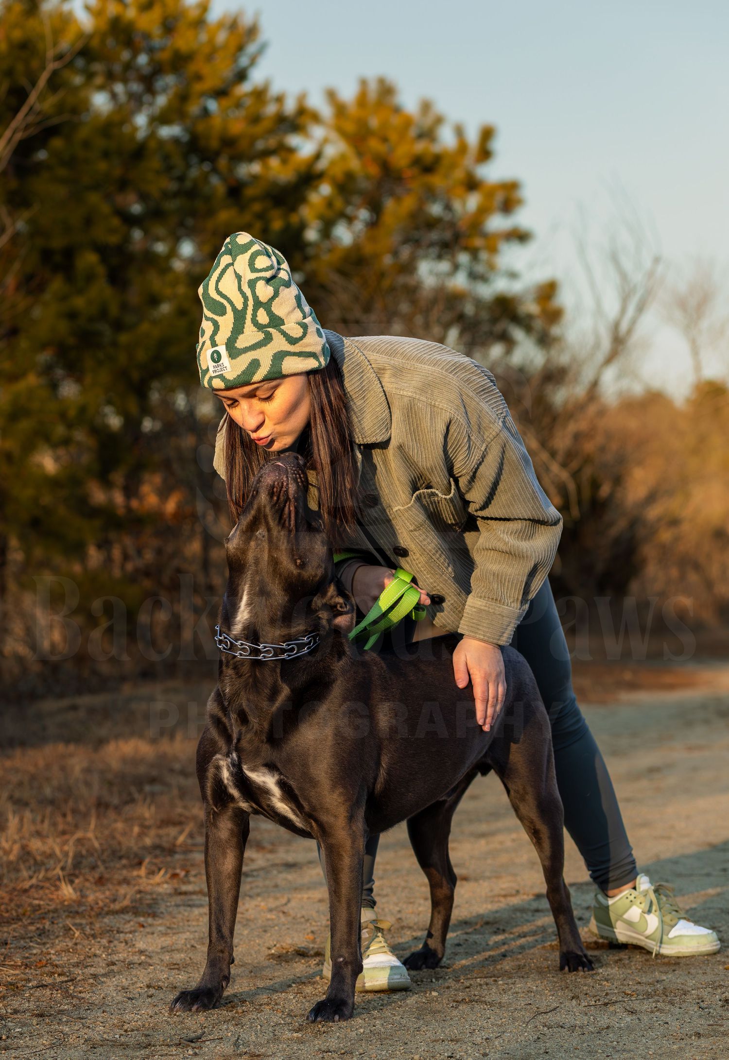 Person in winter hat and jacket bends down to pet a large black dog on a dirt path during autumn.
