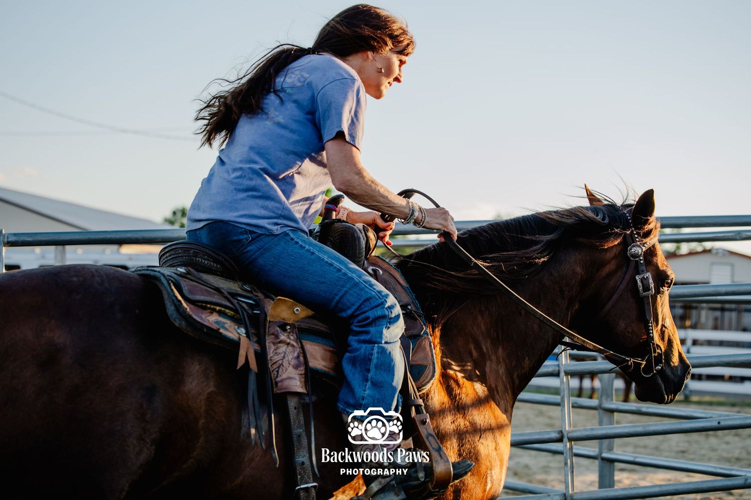 Woman riding a dark bay horse in a rodeo arena, wearing blue jeans and a blue shirt, with hair blowing in the wind.