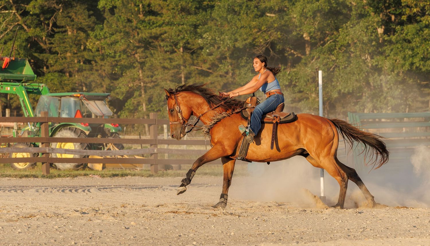 Woman riding a bay horse at a canter in a sandy arena with green tractor and trees in background.