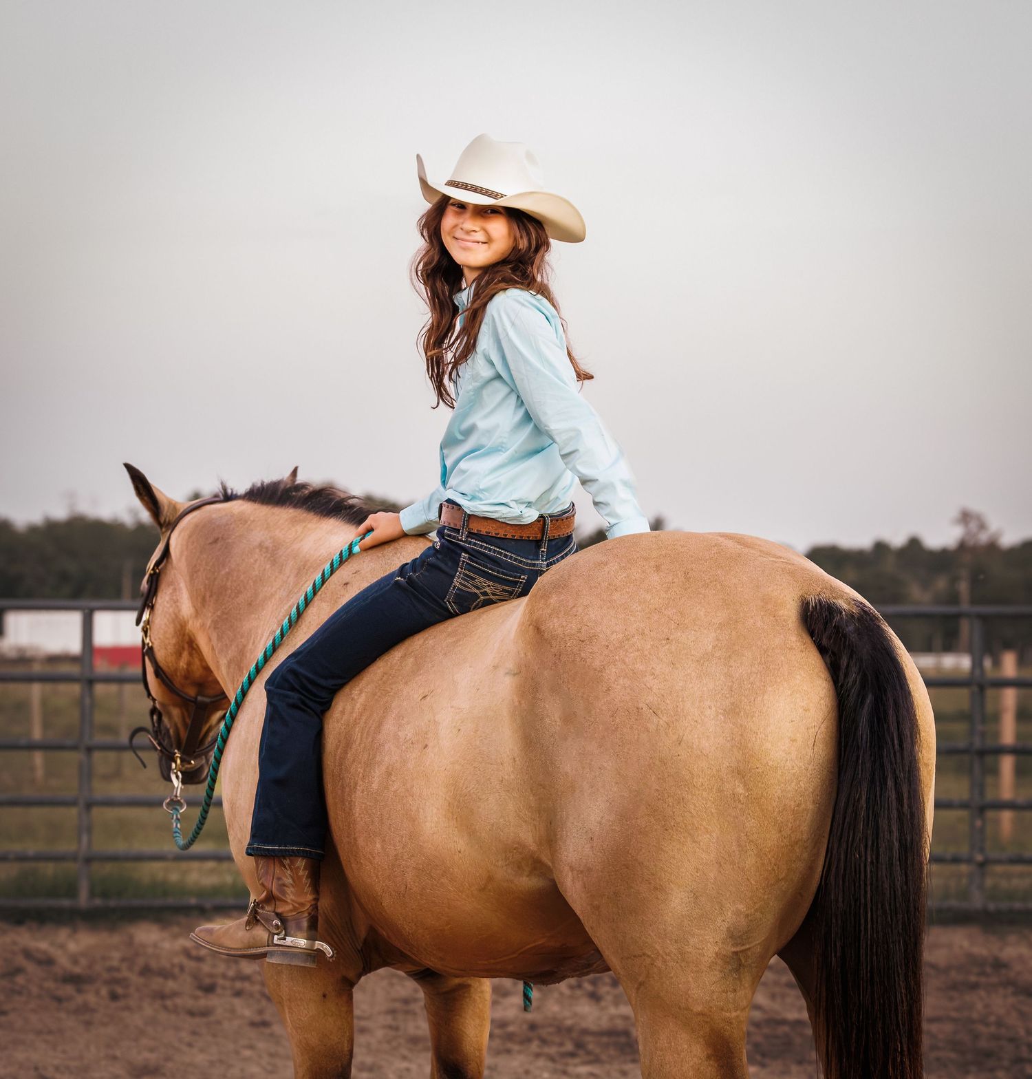 Smiling young woman in cowboy hat rides a buckskin horse in an outdoor arena.
