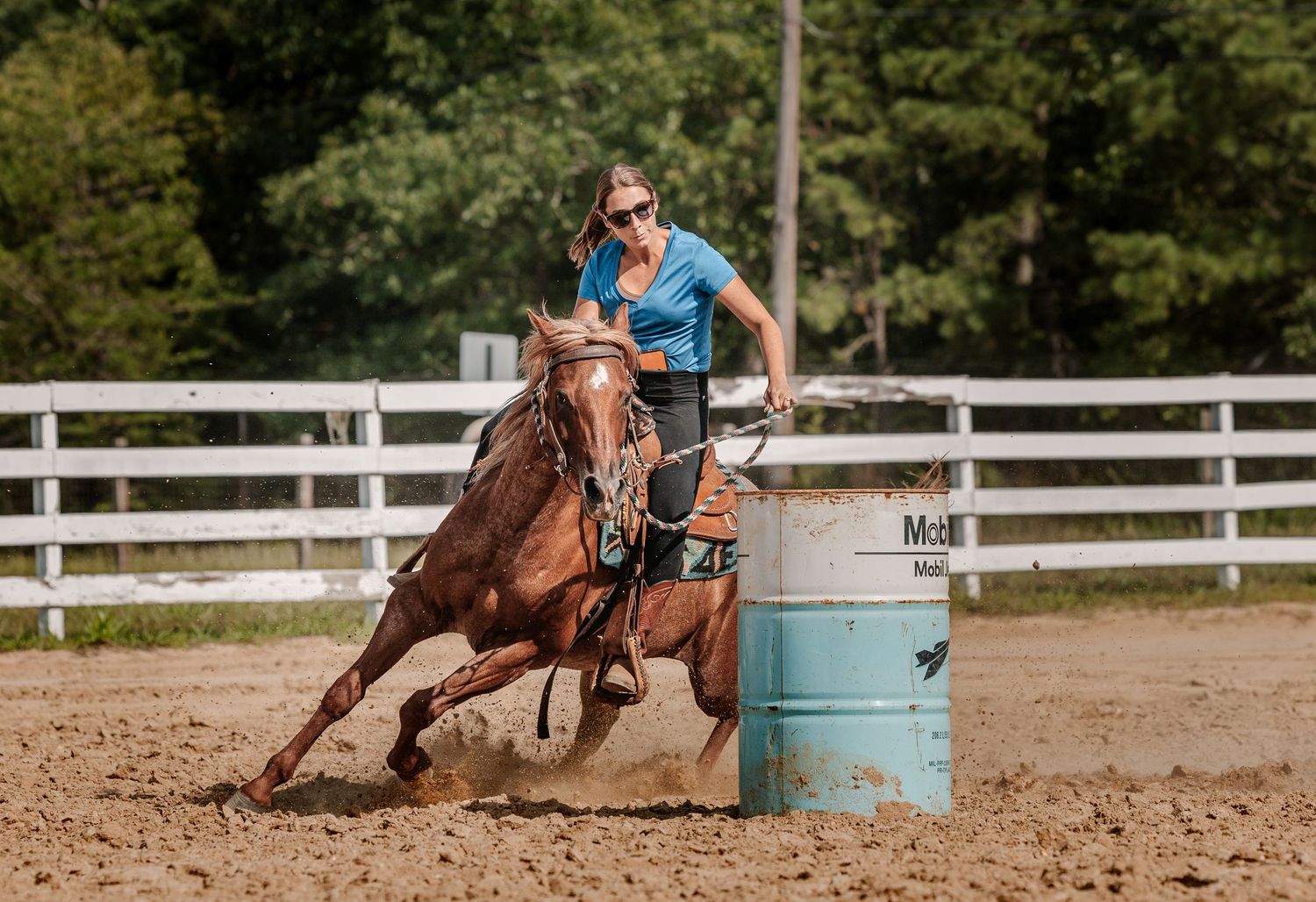 Woman riding chestnut horse around barrel in dirt arena during barrel racing competition.