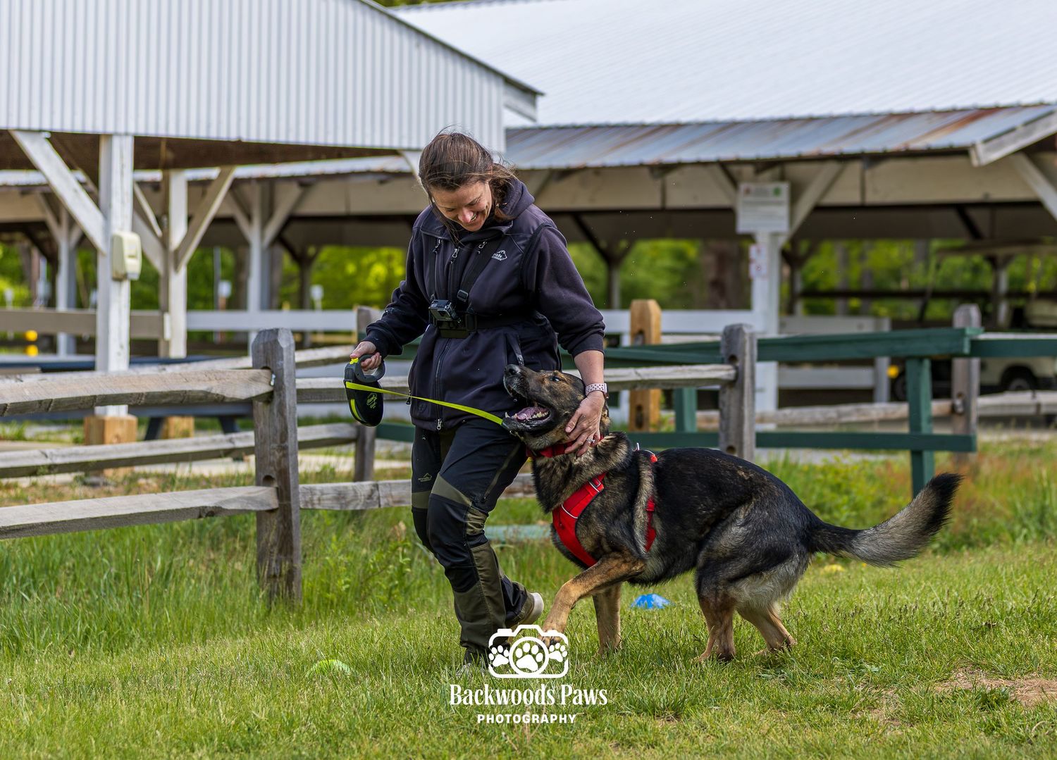Woman in black jacket training a German Shepherd dog on a green lawn near a wooden fence and barn.