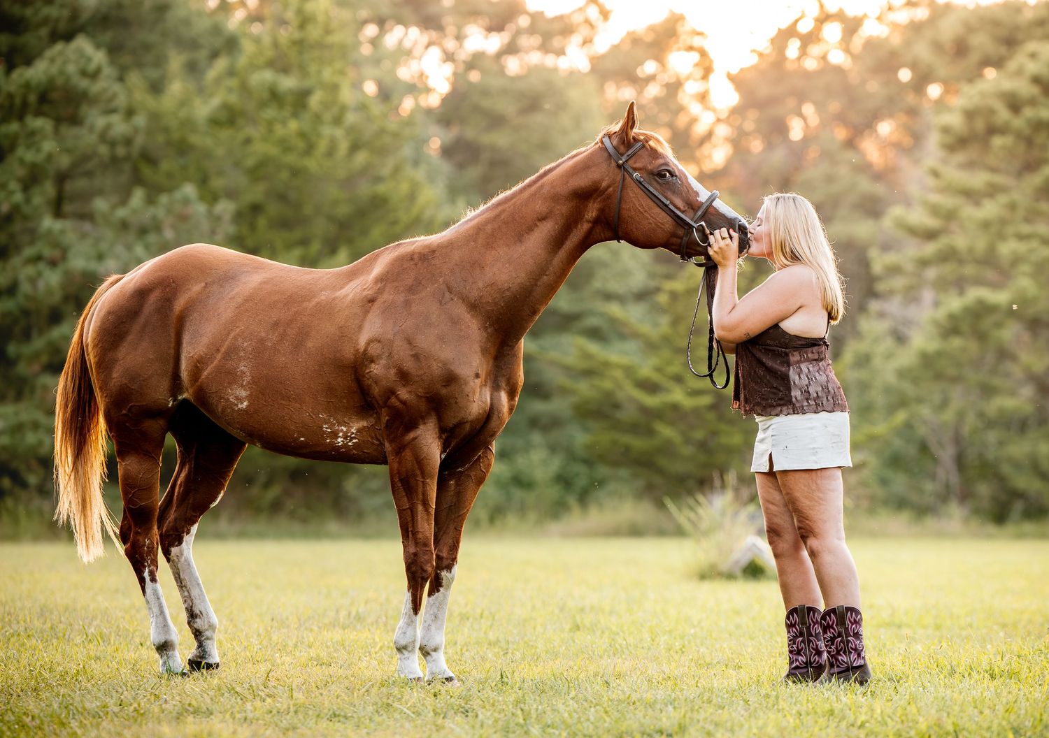 A blonde woman in cowboy boots and shorts affectionately nuzzles a chestnut horse in a sunny green meadow.